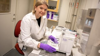 Kvinna i vit labbrock och lila handskar i ett laboratorium.