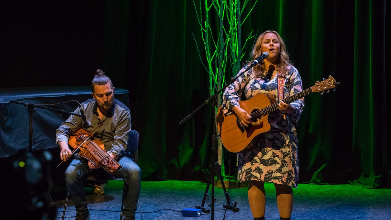 Bild på Carolina Miskovsky som sjunger och spelar gitarr och Daniel Pettersson som spelar nyckelharpa på scenen i Aula Nordica, Umeå universitet.