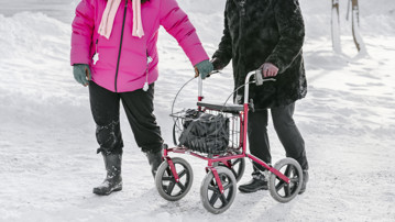Yngre kvinna hjälper äldre med rullator i snö, inga ansikten syns