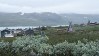 Vy över sjö med kåtor och stugor i förgrunden och fjäll i bakgrunden.