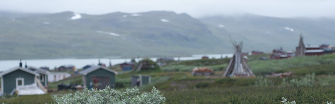 Vy över sjö med kåtor och stugor i förgrunden och fjäll i bakgrunden.