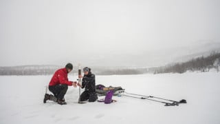 Två forskare på isen på fjällsjö håller på med fältarbete.