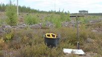 Foto av skogsområde med en hink, arkeologiska provtagningsinstrument och jordborr i förgrunden.