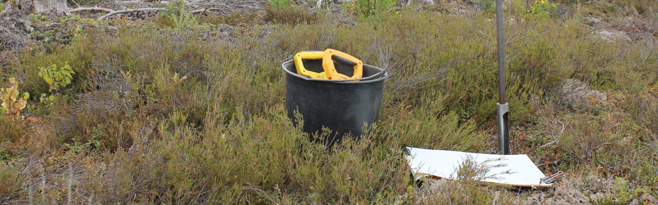 Foto av skogsområde med en hink, arkeologiska provtagningsinstrument och jordborr i förgrunden.