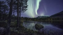 TAIGA är ett centrum i norra Sverige. Bilden visar sjö och skog upplyst av norrsken på natthimmel.