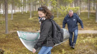 Två personer som bär en kanot genom skogen mot vattnet.