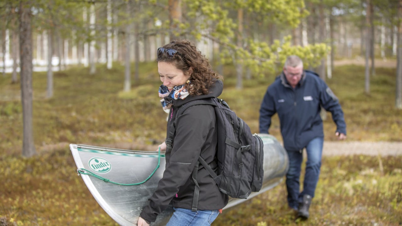Två personer som bär en kanot genom skogen mot vattnet.