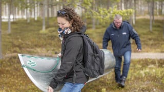 Två personer som bär en kanot genom skogen mot vattnet.