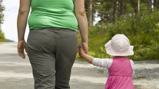 Lätt överviktig mamma går med dotter i handen utomhus, bilden tagen bakifrån