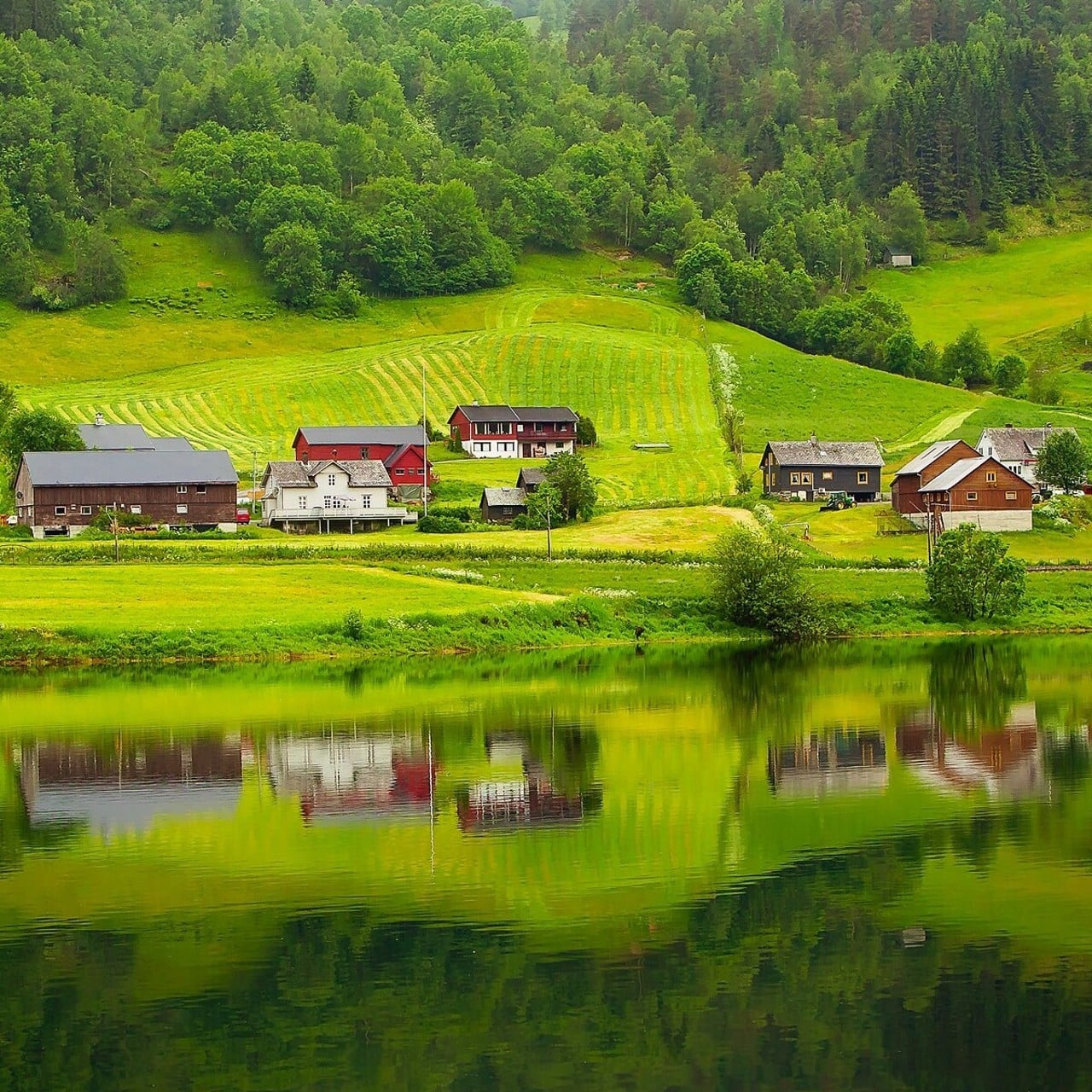 Hus, stugor, bodar och andra byggnader på andra sidan en sjö. Speglingen gör att byggnaderna och de gröna ängarna och bergen bakom speglas i sjöns vatten. Det är sommar och grönt.