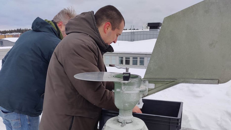 Två personer står på ett hustak framför en apparat i metall