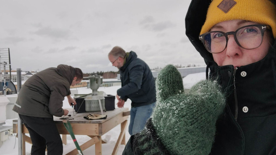 Två personer står vid ett bord på ett hustak böjda över en apparat i metall, en tredje person tittar in i kameran och gör tummen upp.