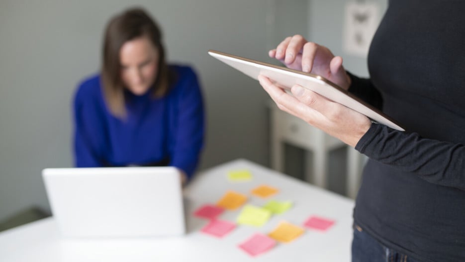Bild på personen som håller laptop och en person i bakgrunden som lägger postitlappar på bordet