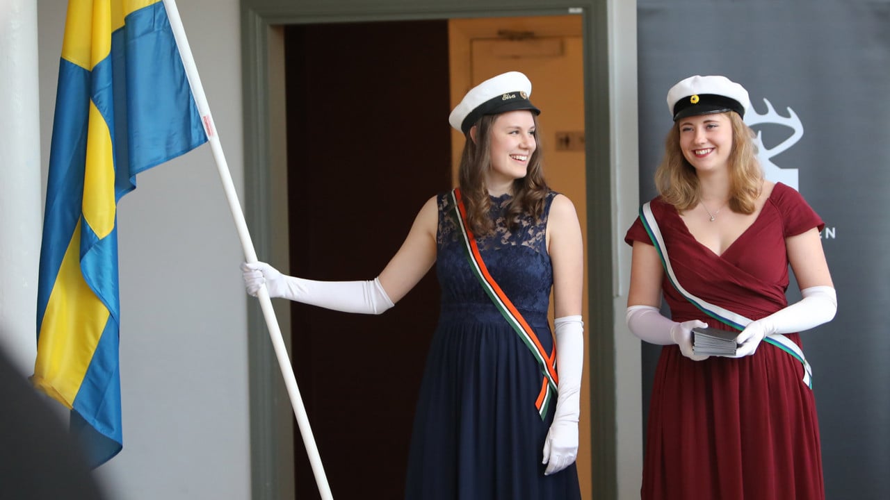 Studentmarskalkar står med flaggor och delar ut program.