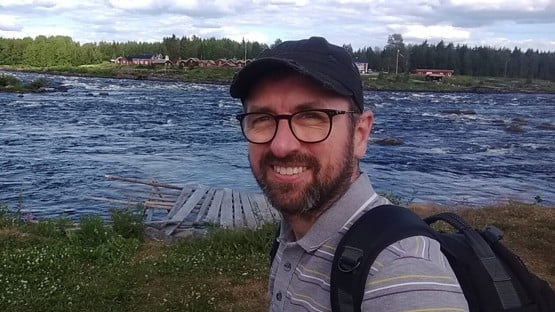 Stuart Wales står framför en älv. Stugor i bakgrunden.