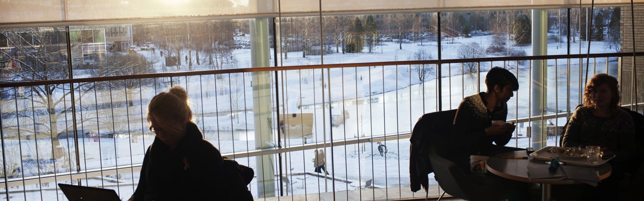 Campus sett från inifrån Lindellhallsfiket en solig vinterdag. Några studenter arbetar med sina datorer.